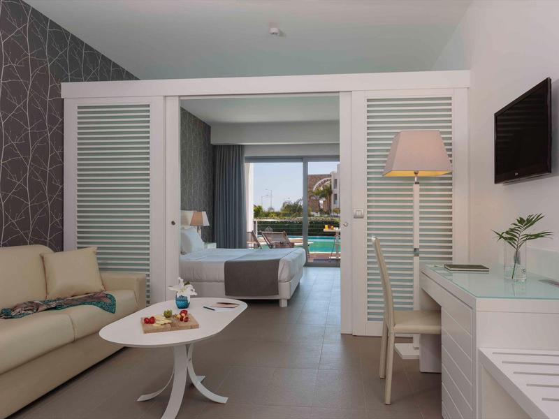 Modern living room with sofa, desk, TV, and view into bedroom overlooking a pool.