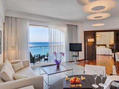 Camera d'hotel luminosa con zona salotto, TV e balcone con vista sul mare.