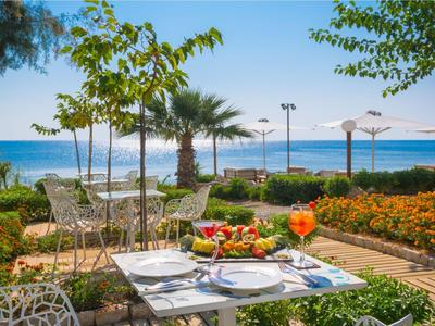 Terrazza con tavolo apparecchiato, vista mare e giardino con palme sotto un cielo sereno.