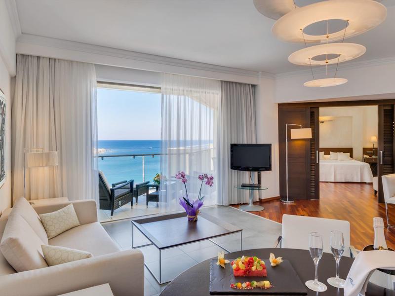 Camera d'hotel luminosa con zona salotto, TV e balcone con vista sul mare.