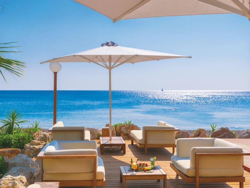 Terrazza con poltrone e ombrellone davanti al mare azzurro sotto il cielo sereno.