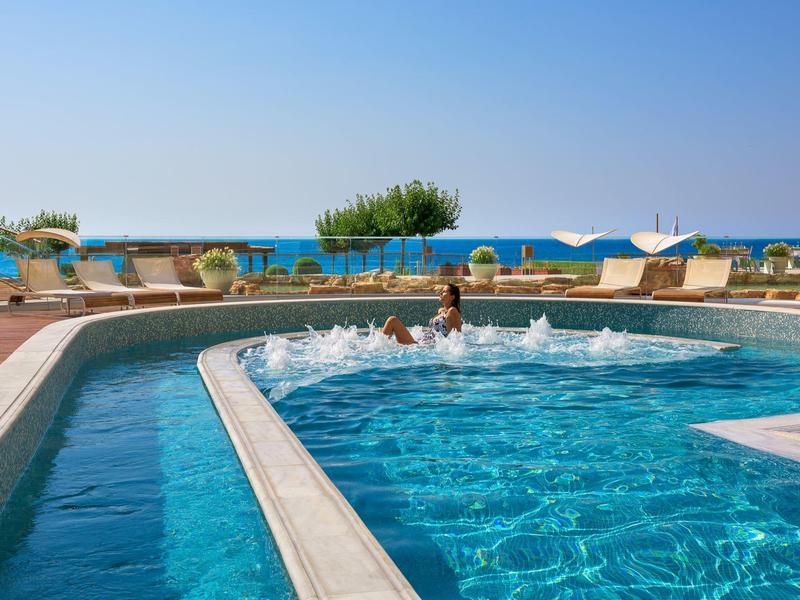 Piscina all'aperto con getti d'acqua, vista sul mare e lettini con ombrelloni.