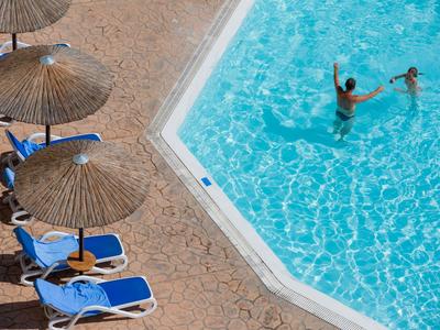 Schwimmbecken mit klarem Wasser, zwei spielenden Kindern und blauen Liegestühlen unter Strohm-Sonnenschirmen.