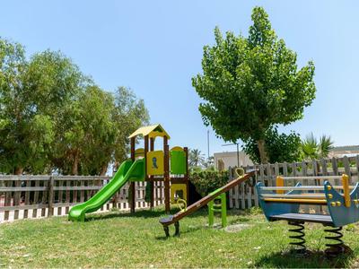 Parco giochi per bambini con struttura per arrampicarsi, scivolo e altalena su prato soleggiato.