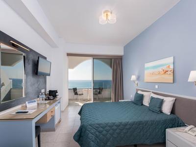 Camera d'hotel con letto blu, TV, scrivania e balcone con vista sul mare.
