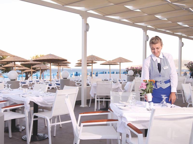 Cameriere prepara il tavolo su una terrazza con sedie bianche e ombrelloni vicino all'acqua.