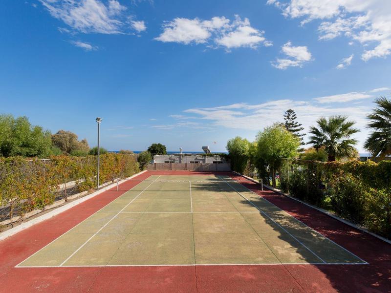 Campo da tennis vuoto circondato da cespugli e palme sotto un cielo azzurro.