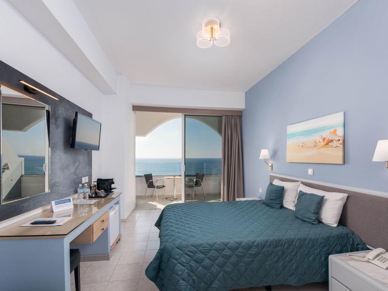 Camera d'hotel con letto blu, TV, scrivania e balcone con vista sul mare.