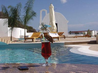 Szklanka z koktajlem przy basenie z białymi budynkami, leżakami i roślinami w tle.