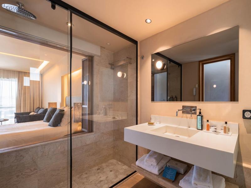 Moderne hotelbadkamer met glazen douche, grote spiegel en open zicht naar slaapkamer.