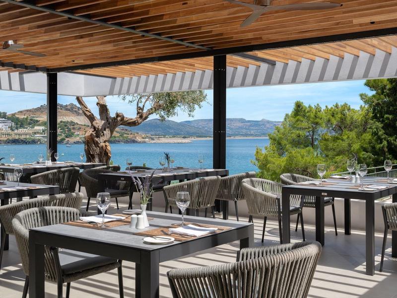Open restaurant met moderne tafels en stoelen met uitzicht op zee en kustlijn.