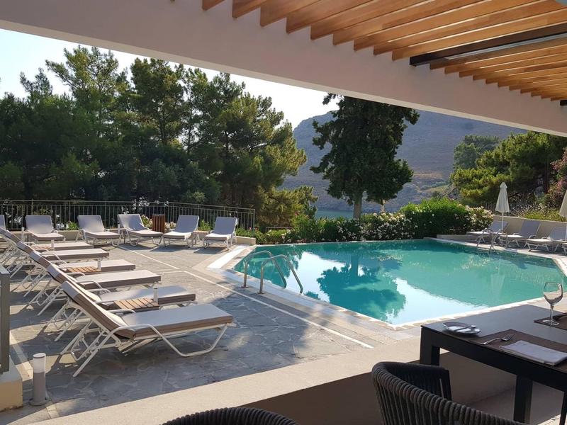 Ligstoelen bij het zwembad onder een houten pergola met groene bomen en bergen op de achtergrond.