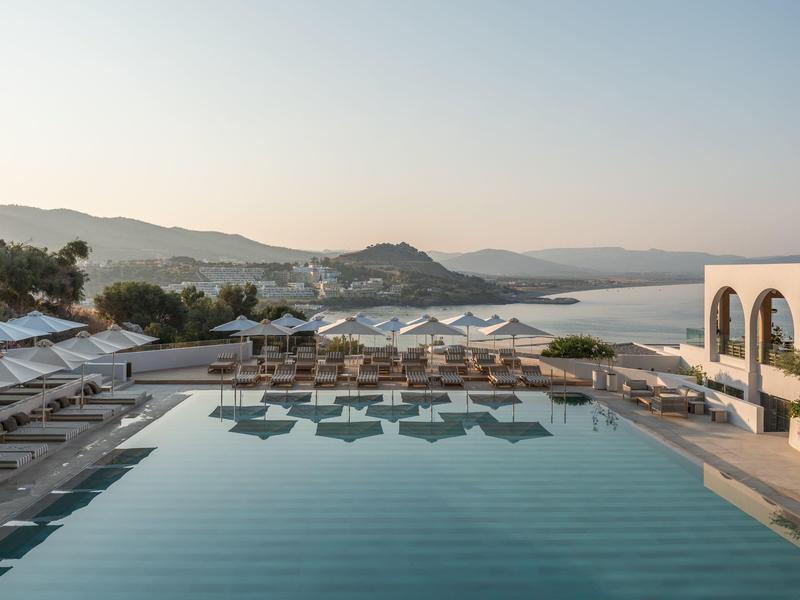 Zwembad met ligstoelen en parasols met uitzicht op bergen en meer bij zonsondergang.