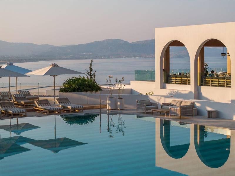 Een rustige zwembadzone met ligstoelen en parasols met uitzicht op zee en heuvels.