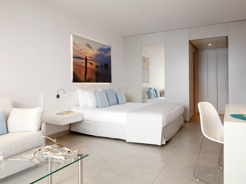 Heldere moderne slaapkamer met wit interieur, tweepersoonsbed, bank en glazen salontafel.