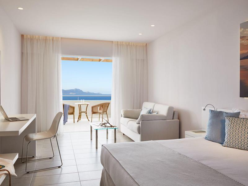 Lichte hotelkamer met zeezicht, bed, bank, bureau en balkon met stoelen.