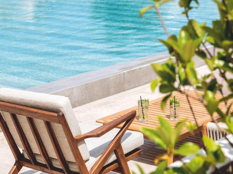 Houten ligstoel met kussen aan zwembadrand en planten op de voorgrond, ontspannen vakantiesfeer