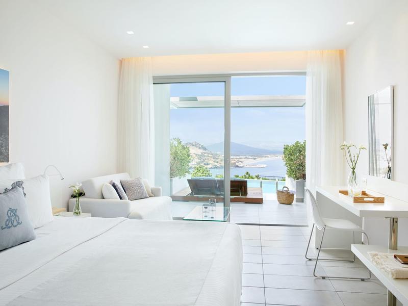 Lichte slaapkamer met bed, bank, bureau en grote schuifdeur met uitzicht op zee.