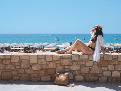 Mujer sentada relajada en un muro de piedra con vista a la playa y el mar en un día soleado.