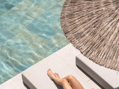 Frau entspannt am Pool auf Liege unter Sonnenschirm mit Wasser im Hintergrund.
