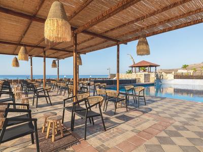 Offene Terrasse mit Holzdach, Stühlen, Tischen und Blick auf einen Pool und das Meer im Hintergrund.