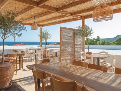 Hell gestaltete Terrasse mit Holztischen, Stühlen, Pflanzen und Blick auf das blaue Meer.