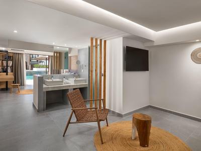 Moderner Wohnraum mit grauem Boden, Holzstuhl, Teppich, TV an der Wand und Küchenbereich im Hintergrund.