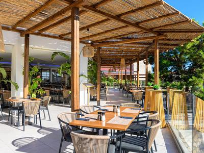 Offene Terrasse eines Hotels mit Tischen, Stühlen und Pflanzen unter einem Holzpergola.