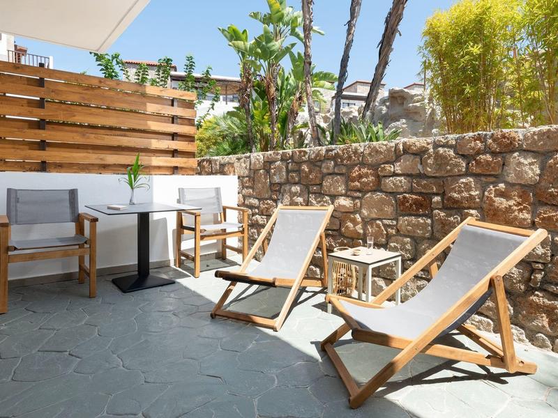 Terrazza soleggiata con due sdraio in legno e un piccolo tavolo con sedie davanti a un muro di pietra.