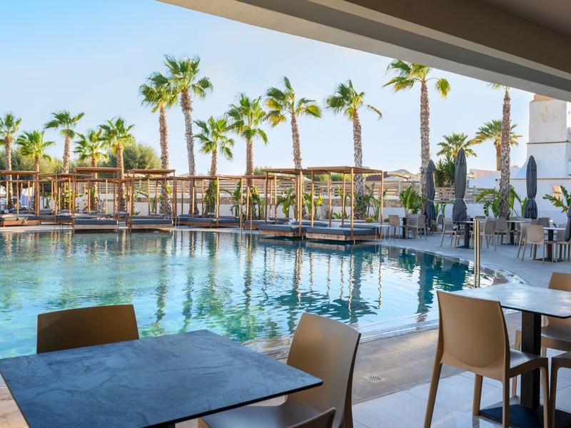 Vista di una piscina moderna con sedie e tavoli sotto un'area coperta e palme sullo sfondo.