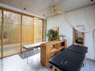 Hell eingerichteter Raum mit Massageliegen, Holzregal, Pflanzen und großen Fenstern mit Bambusjalousien.