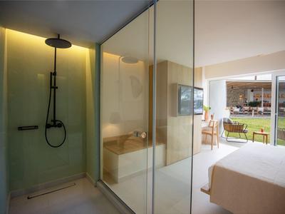 Modernes Schlafzimmer mit Glasdusche, beigem Bett und Blick auf Terrasse mit Rasenfläche.