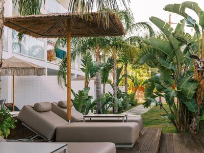 Outdoor-Liegestühle mit Strohdach im tropischen Garten neben weißem Gebäude und grünen Pflanzen.