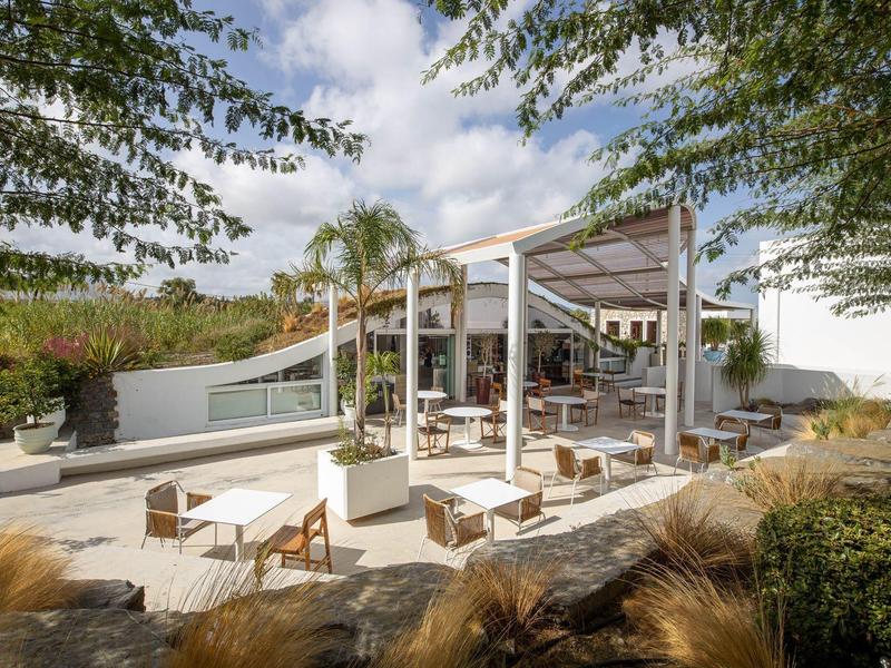 Area esterna moderna di un hotel con pergolato, sedie e tavoli circondati da piante.