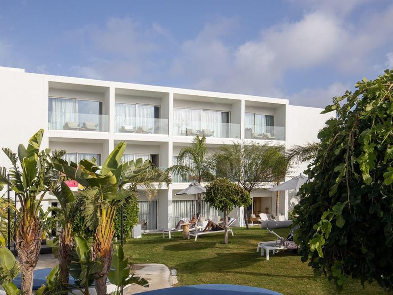 Edificio d'hotel bianco moderno con balconi, circondato da piante verdi e cielo azzurro.