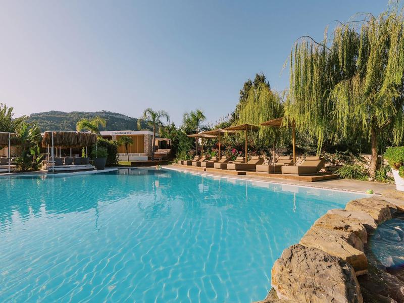 Piscina dell'hotel circondata da lettini e vegetazione sotto un cielo limpido.