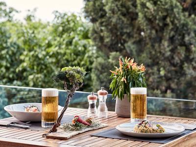 Stół na balkonie z jedzeniem, dwoma szklankami piwa i widokiem na zielone drzewa.