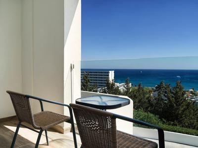 Balcone con sedie e tavolo con vista sul mare e alberi verdi sotto un cielo azzurro.