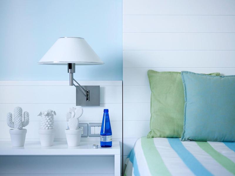 Camera luminosa con letto verde-blu, cuscini e tre piante di cactus bianche su una mensola.