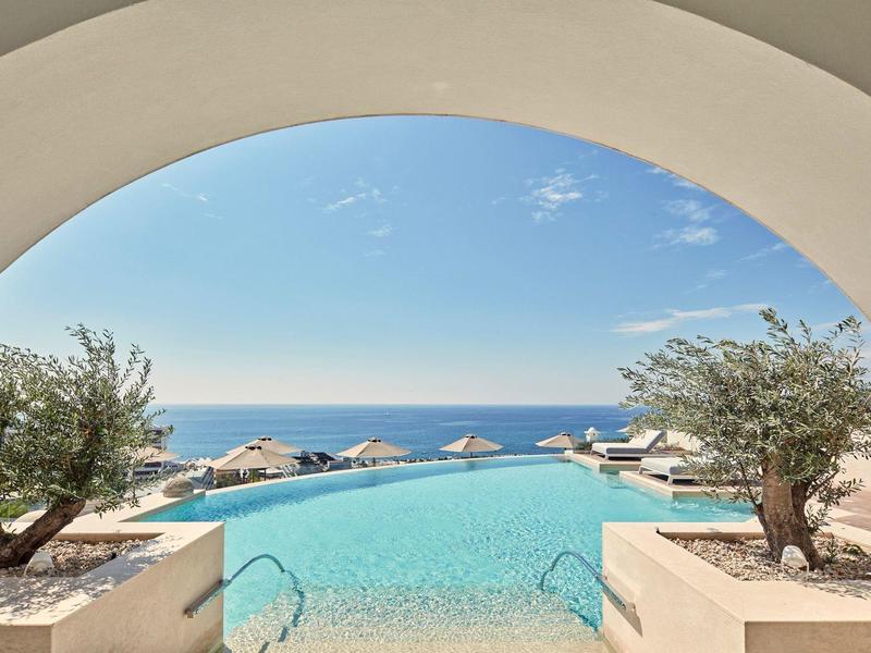 Piscina a sfioro con vista sul mare, circondata da lettini e ulivi sotto un cielo azzurro.