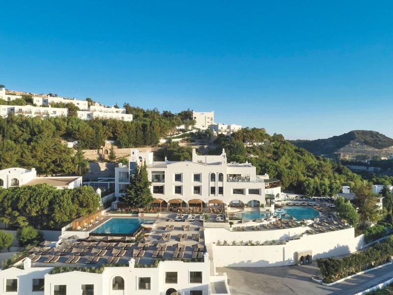 Complesso alberghiero con piscine e edifici bianchi su una collina sotto un cielo blu chiaro.