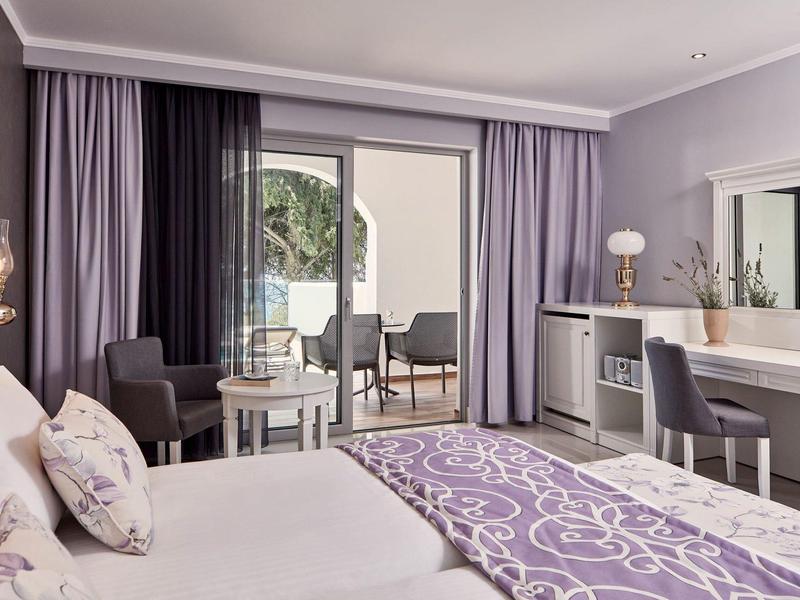 Camera d'albergo luminosa con letto matrimoniale, tende viola e balcone con vista piscina.