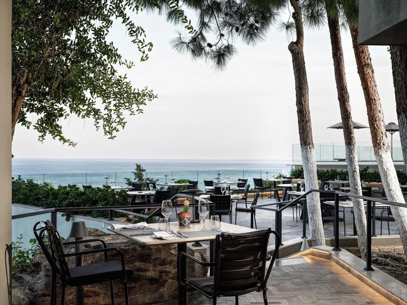 Terrazza di un ristorante con vista sul mare, tavoli apparecchiati e sedie nere sotto gli alberi.