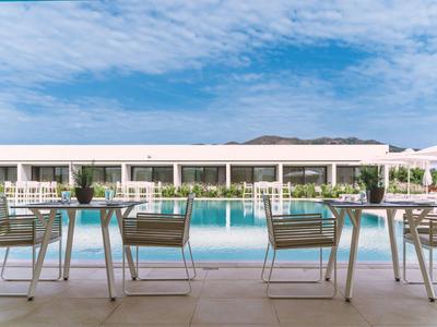 Blick auf einen Poolbereich mit Tischen und Stühlen vor einem modernen Hotelgebäude bei klarem Himmel.