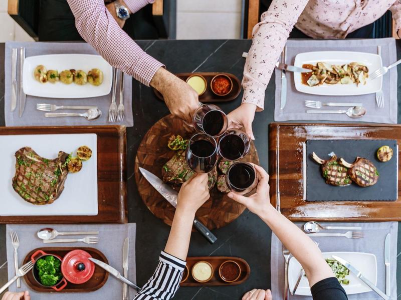 Persone che brindano con bicchieri di vino sopra un tavolo apparecchiato con piatti di carne e verdure.