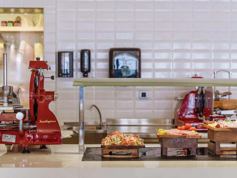 Area buffet con pane, affettati e due affettatrici rosse davanti a una parete piastrellata bianca.