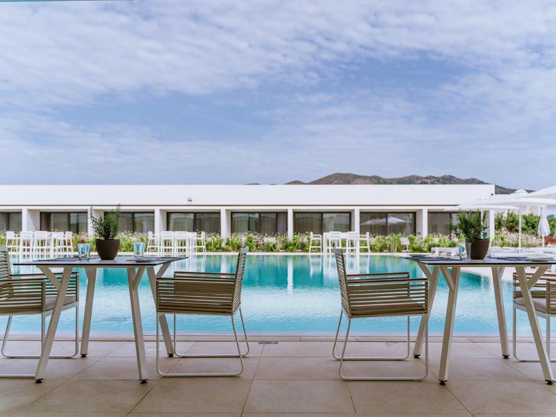 Piscina all'aperto moderna con area salotto e lettini sotto un cielo limpido accanto a un hotel.