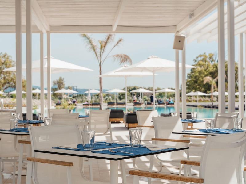 Area ristorante all'aperto moderna con arredi bianchi vista piscina e palme.