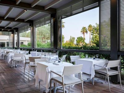 Modernes, offenes Restaurant mit weißen Tischen und Stühlen, großem Fenster und Blick auf Palmen.