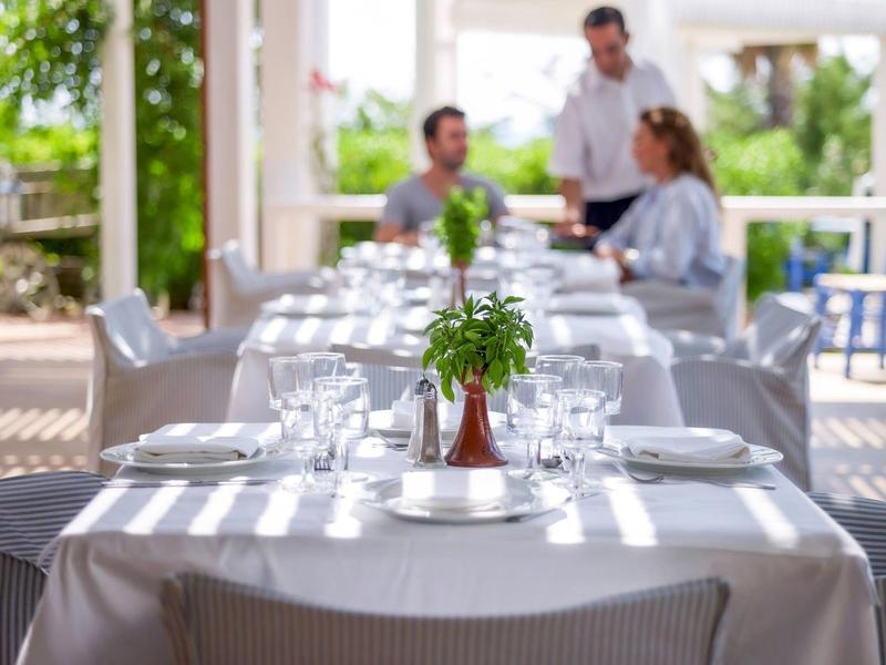 Tavolo apparecchiato con tovaglia bianca e erbe, cameriere che serve sullo sfondo.